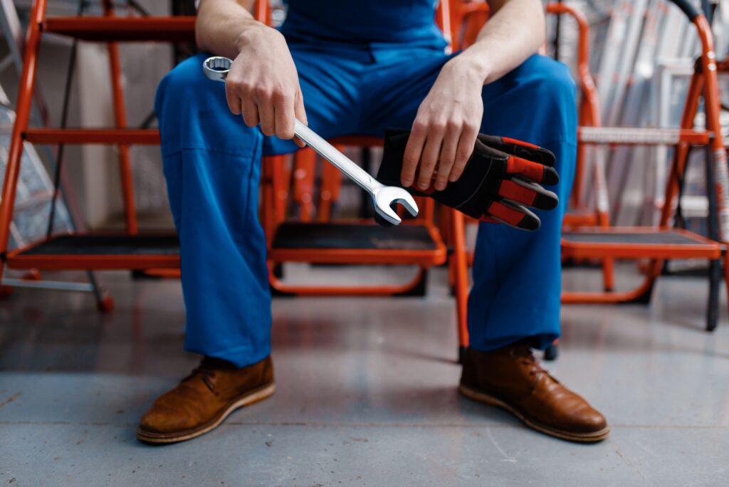 Ein Handwerker in blauer Arbeitskleidung hält einen Schraubenschlüssel und Arbeitshandschuhe, bereit für die Arbeit. Im Hintergrund sind Leitern und Werkzeuge zu sehen