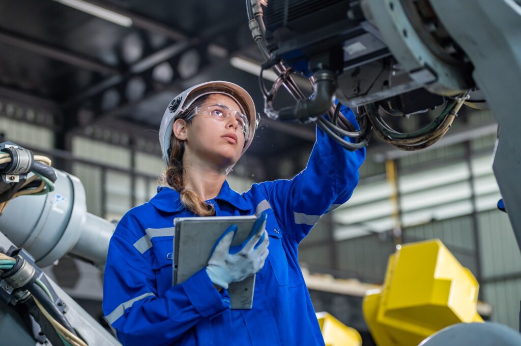 Eine Ingenieurin in blauer Schutzkleidung und Helm inspiziert eine industrielle Montagevorrichtung. Sie hält ein Tablet und trägt Schutzbrille, während sie in einer Fabrikumgebung arbeitet